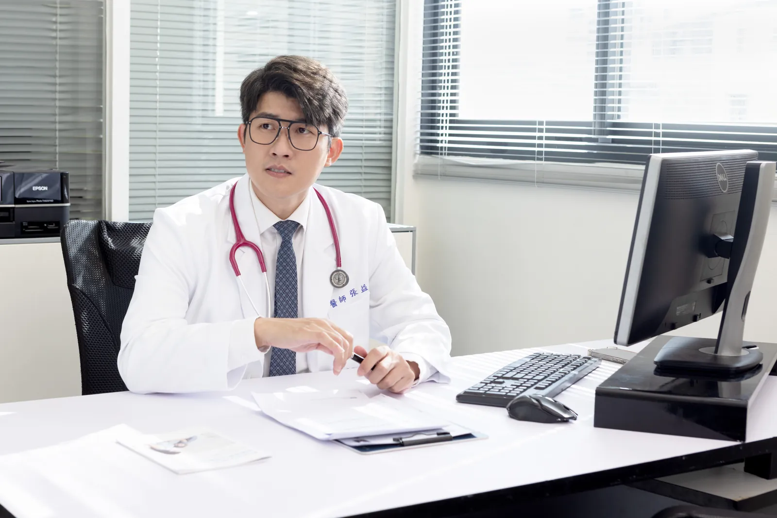 張益豪醫師身穿白袍、掛著聽診器坐在診療桌前，手持筆正在說明，神情專注