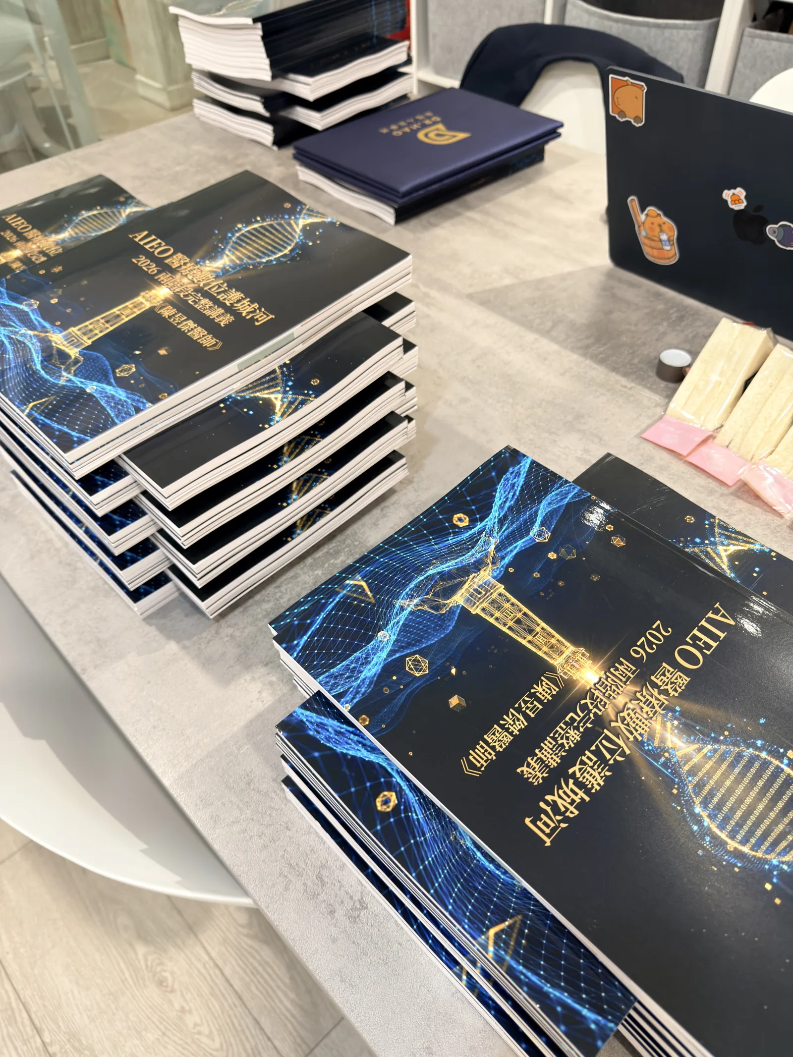 桌面上堆疊的深藍色講義，封面印著 AIEO 醫療數位護城河 2026 兩階段完整講義與陳昱傑醫師字樣，封面有燙金的 DNA 螺旋與燈塔圖案