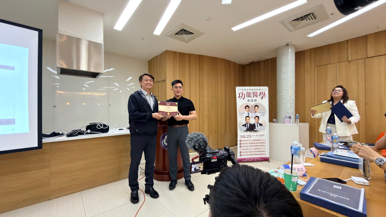 兩人在講台前持文件合影，旁邊一位參與者手持資料夾。
