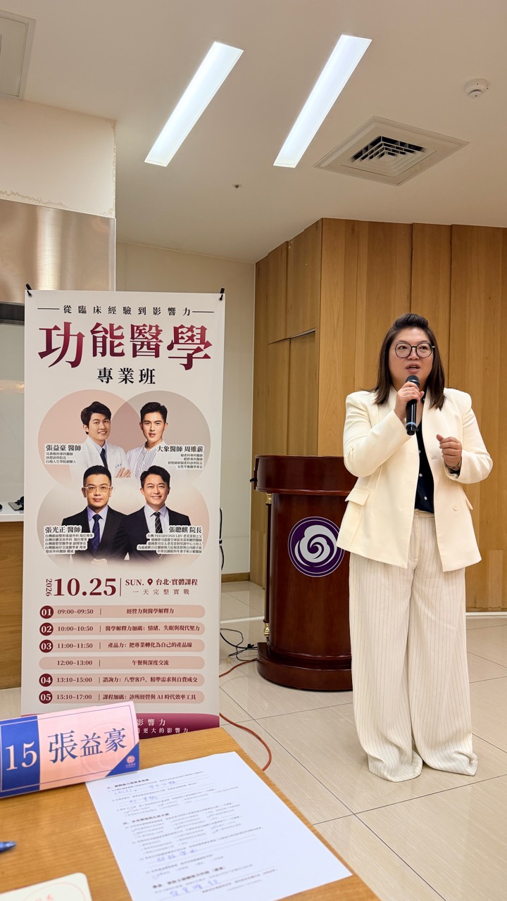 一位穿白色套裝的發言者在課程海報旁持麥克風分享。