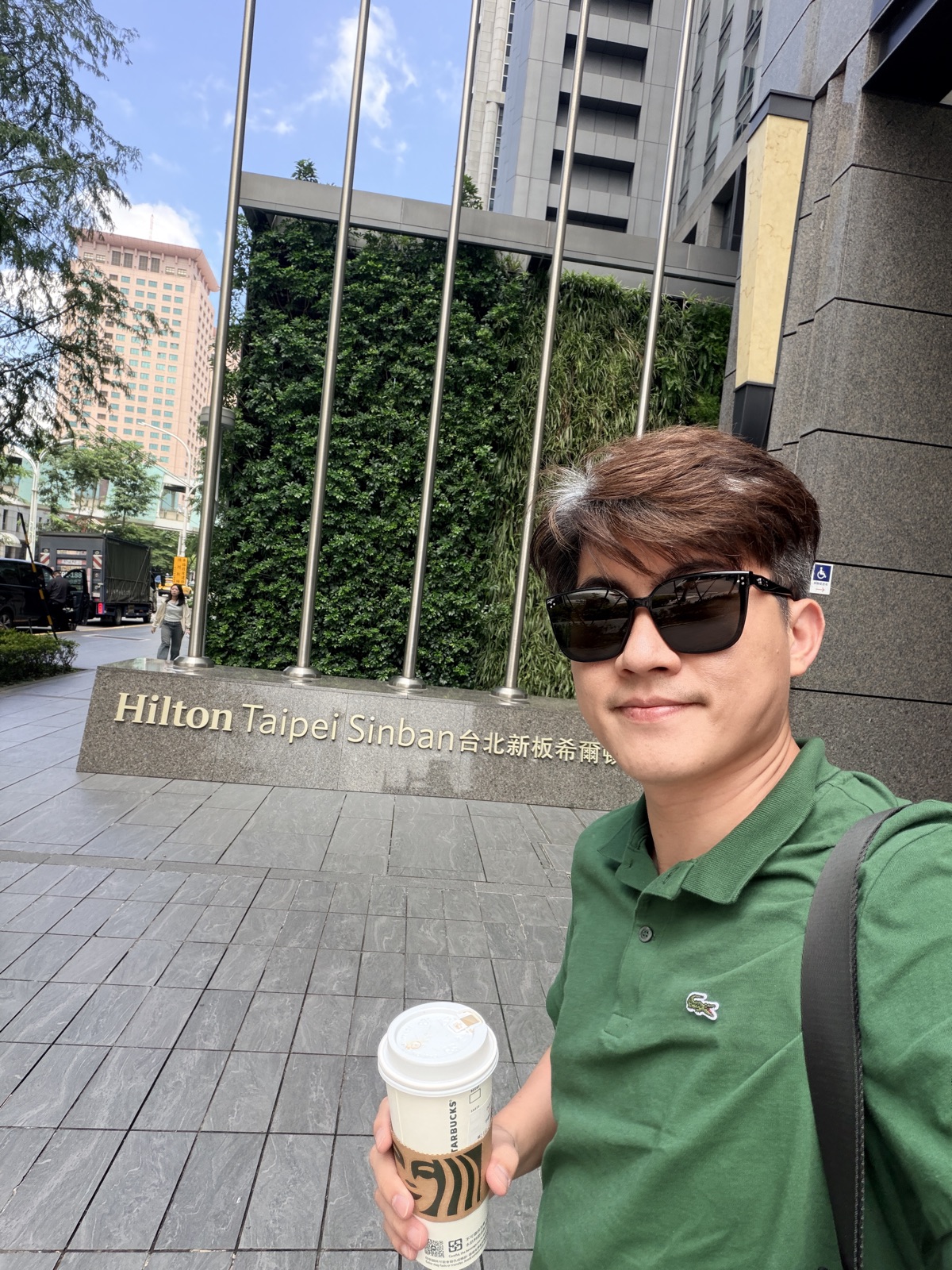 張益豪醫師抵達台北新板希爾頓酒店門口自拍，身後是飯店招牌與綠色植生牆，手拿咖啡準備參加台灣醫美健康管理學會研討會