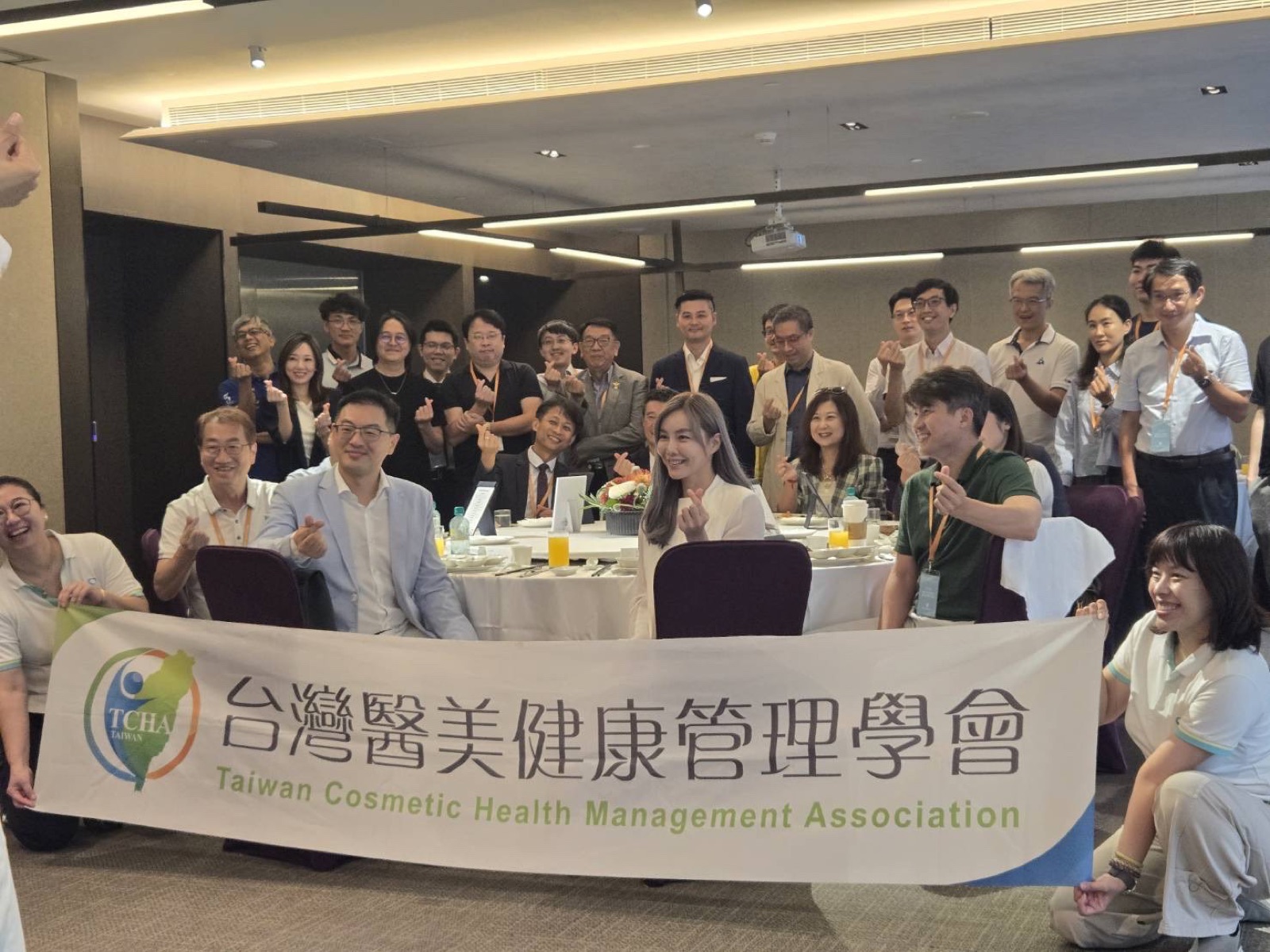 台灣醫美健康管理學會理監事交接暨醫學新知研討會全體與會者大合照，眾人在會議廳內比出愛心手勢，前排拉著印有學會中英文名稱的白色橫幅