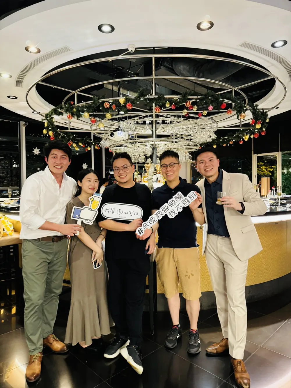 五位與會者在圓形吧台前合影，其中一位舉著手舉牌