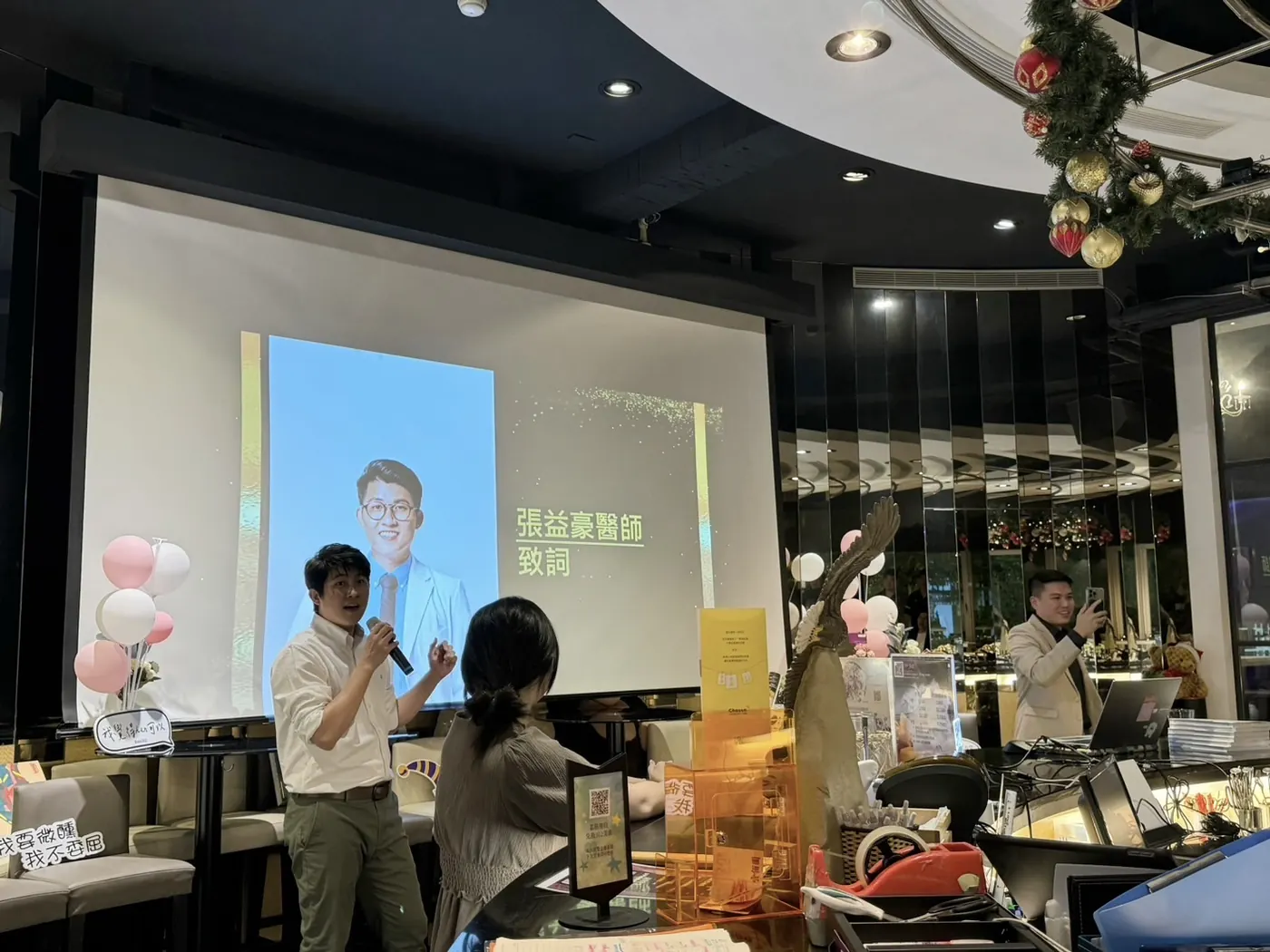張益豪醫師手持麥克風，站在投影幕「張益豪醫師致詞」頁前