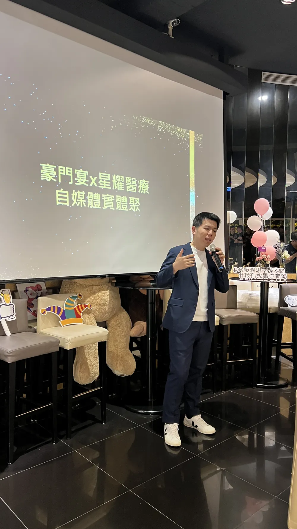 一位穿深藍色西裝的與會者在投影幕前手持麥克風發言