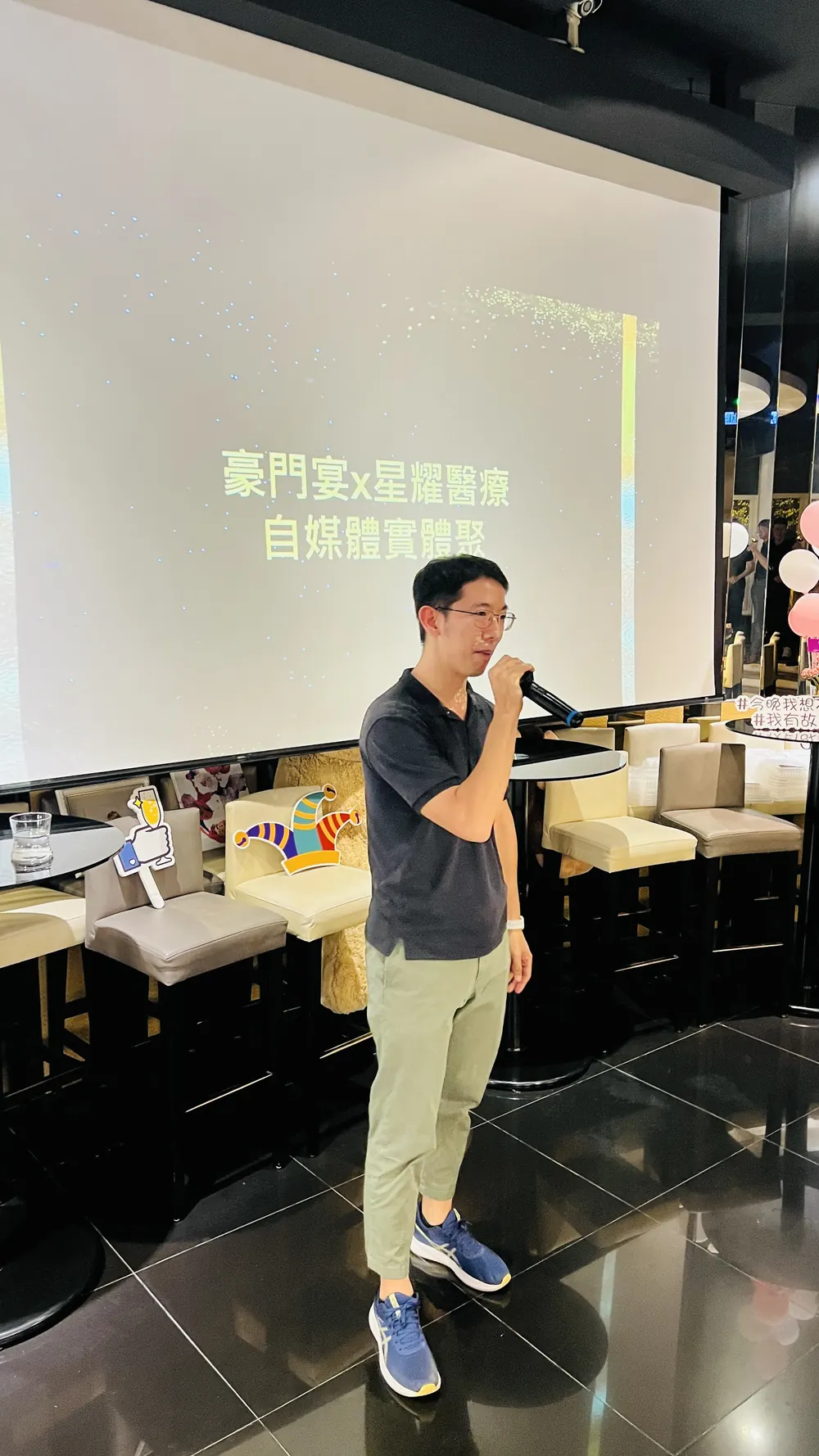 一位穿深色上衣與淺綠色長褲的與會者在投影幕前手持麥克風