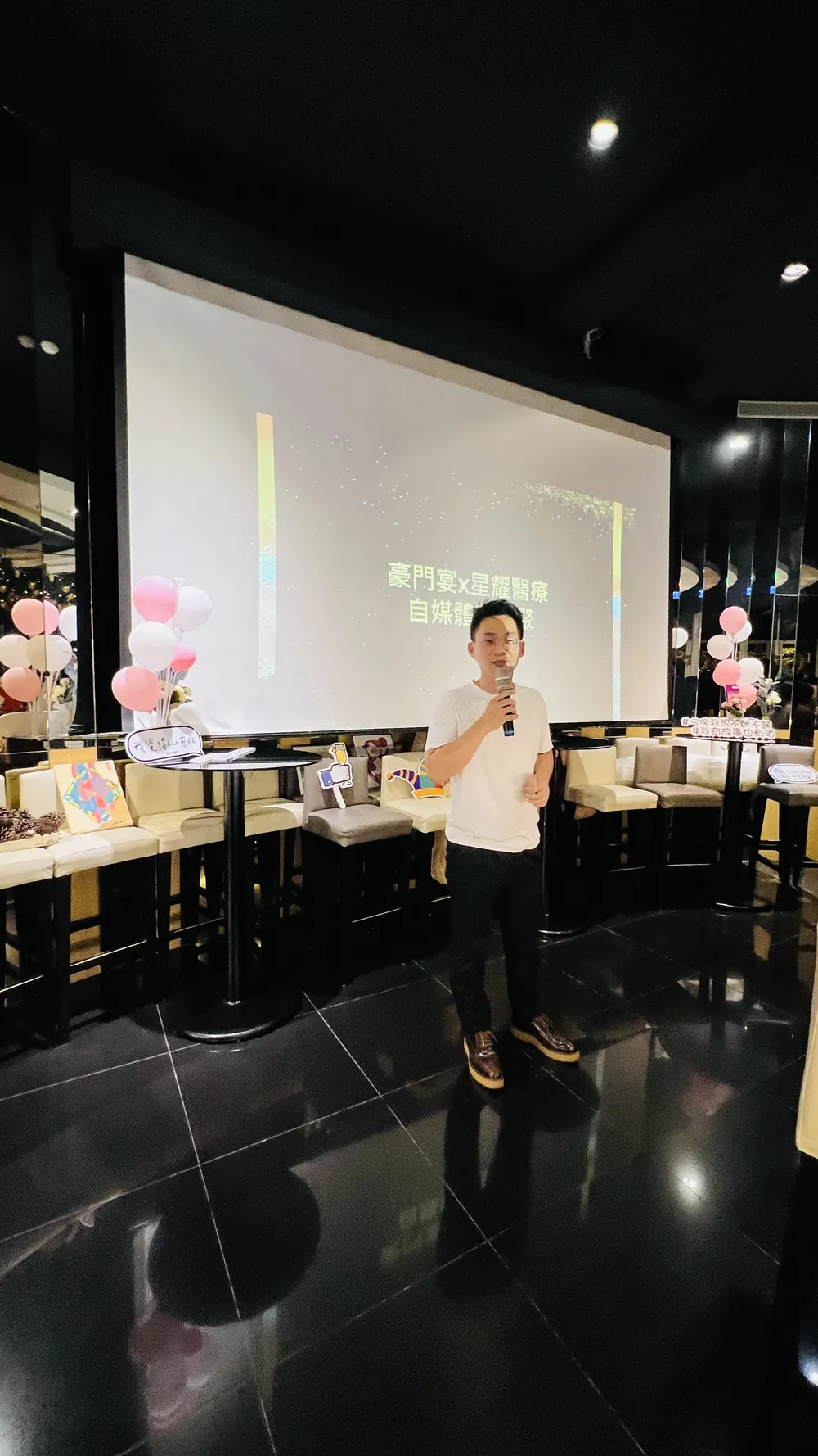 遠景：一位穿白色上衣的與會者站在投影幕前發言，前方是吧台座位