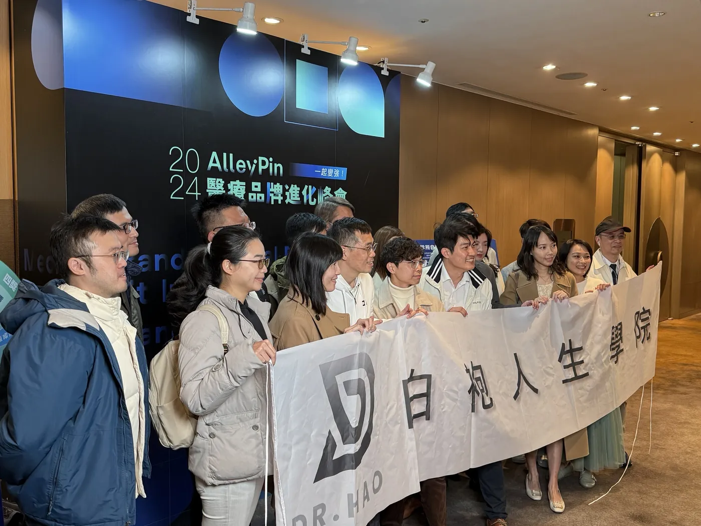 另一個角度的入口合照：成員們靠近鏡頭，拉著「白袍人生學院」布條站在峰會主視覺背板前