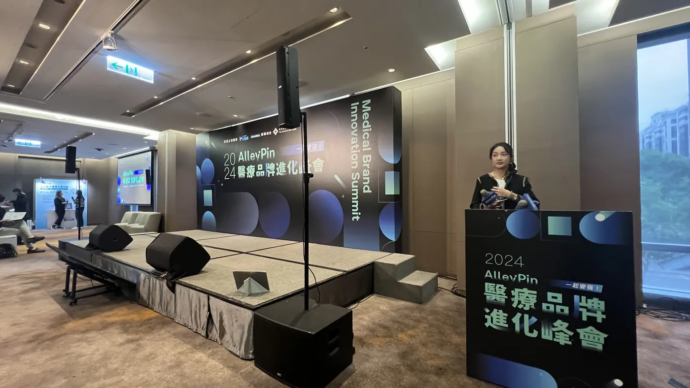會場全景：架高的舞台靠牆，後方立著峰會主視覺背板，右側講台前站著一個人