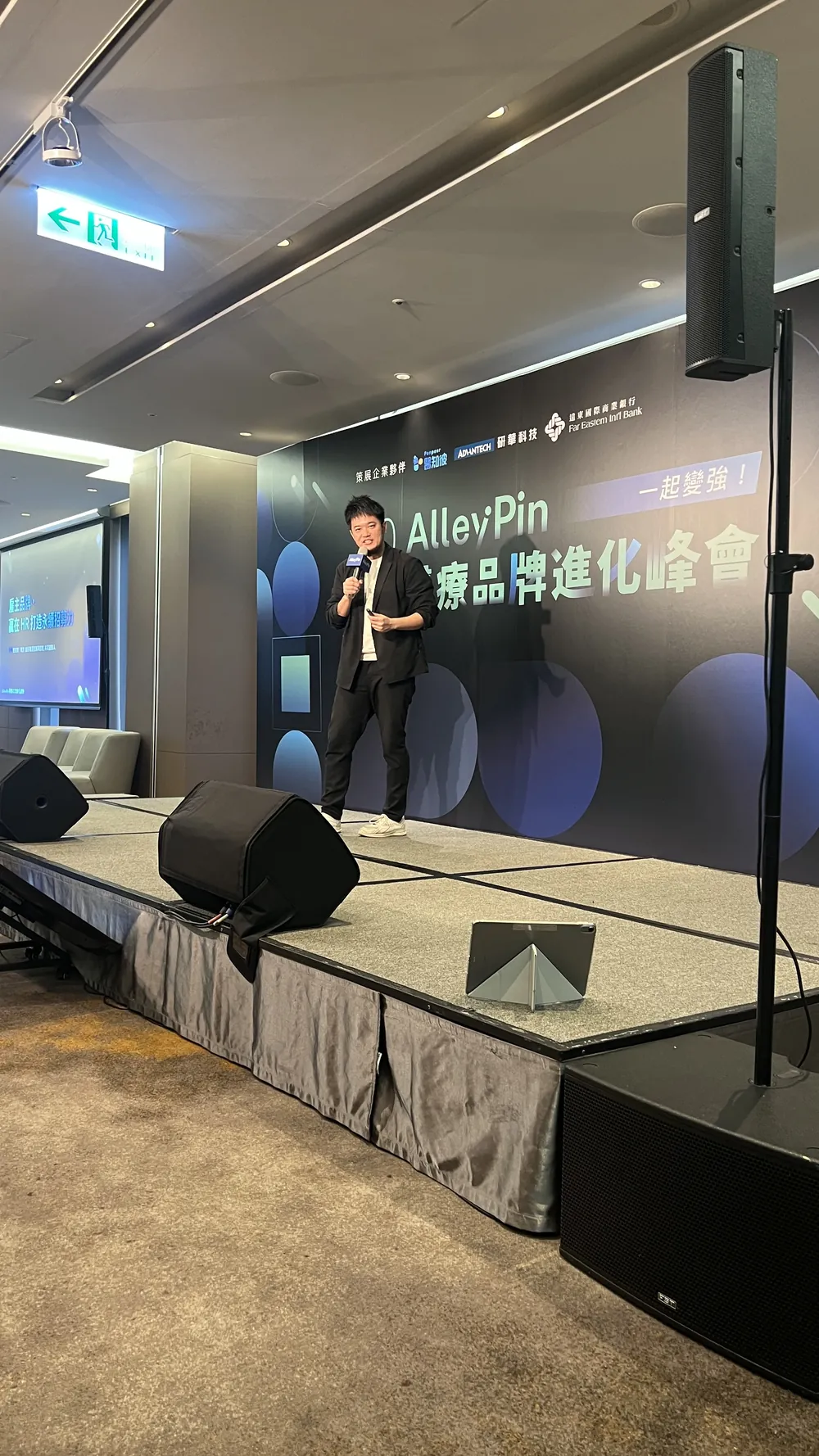 直式拍攝：一位講者手持麥克風站在舞台上分享，後方是峰會主視覺背板
