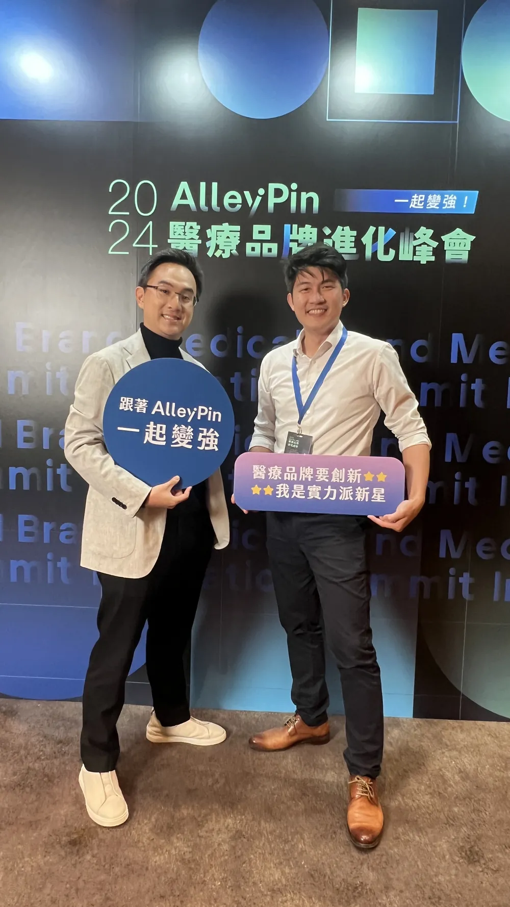 張益豪醫師與一位與會者在峰會主視覺背板前，各自手持 AlleyPin 的標語牌：圓形牌寫「跟著 AlleyPin 一起變強」，長方牌寫「醫療品牌要創新，我是實力派新星」