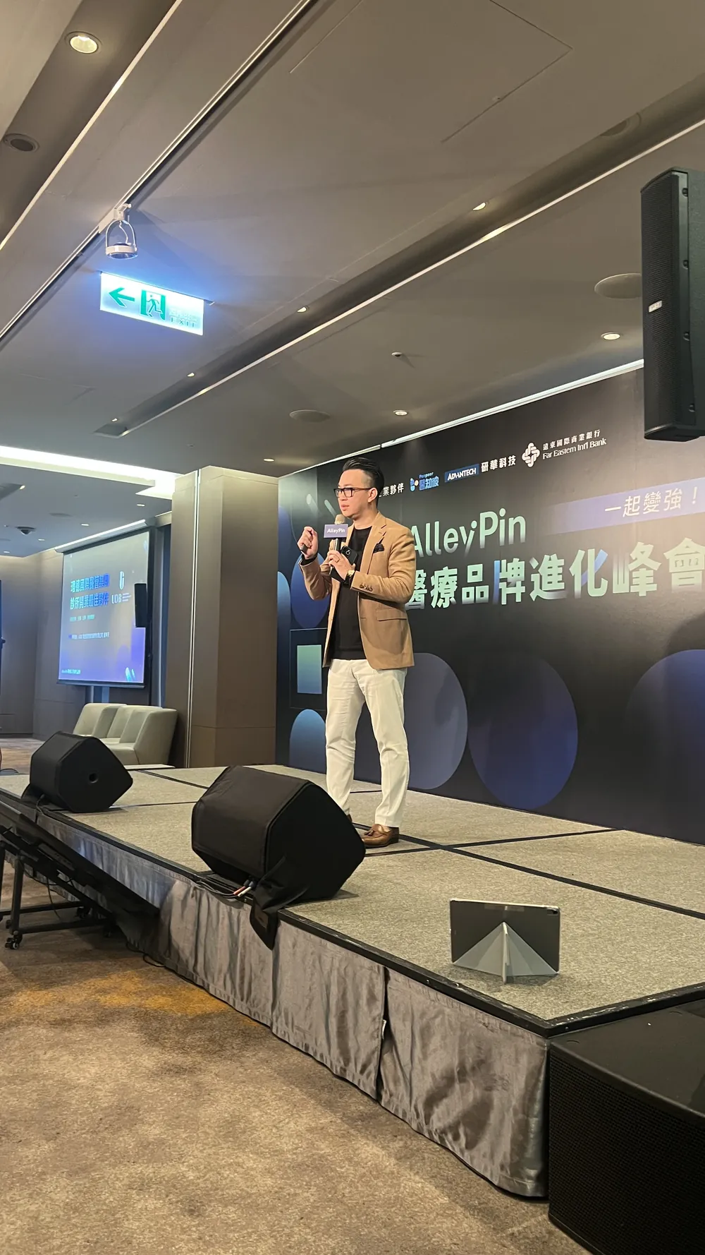 直式拍攝：一位穿米色外套的講者手持麥克風站在舞台上，後方是峰會背板