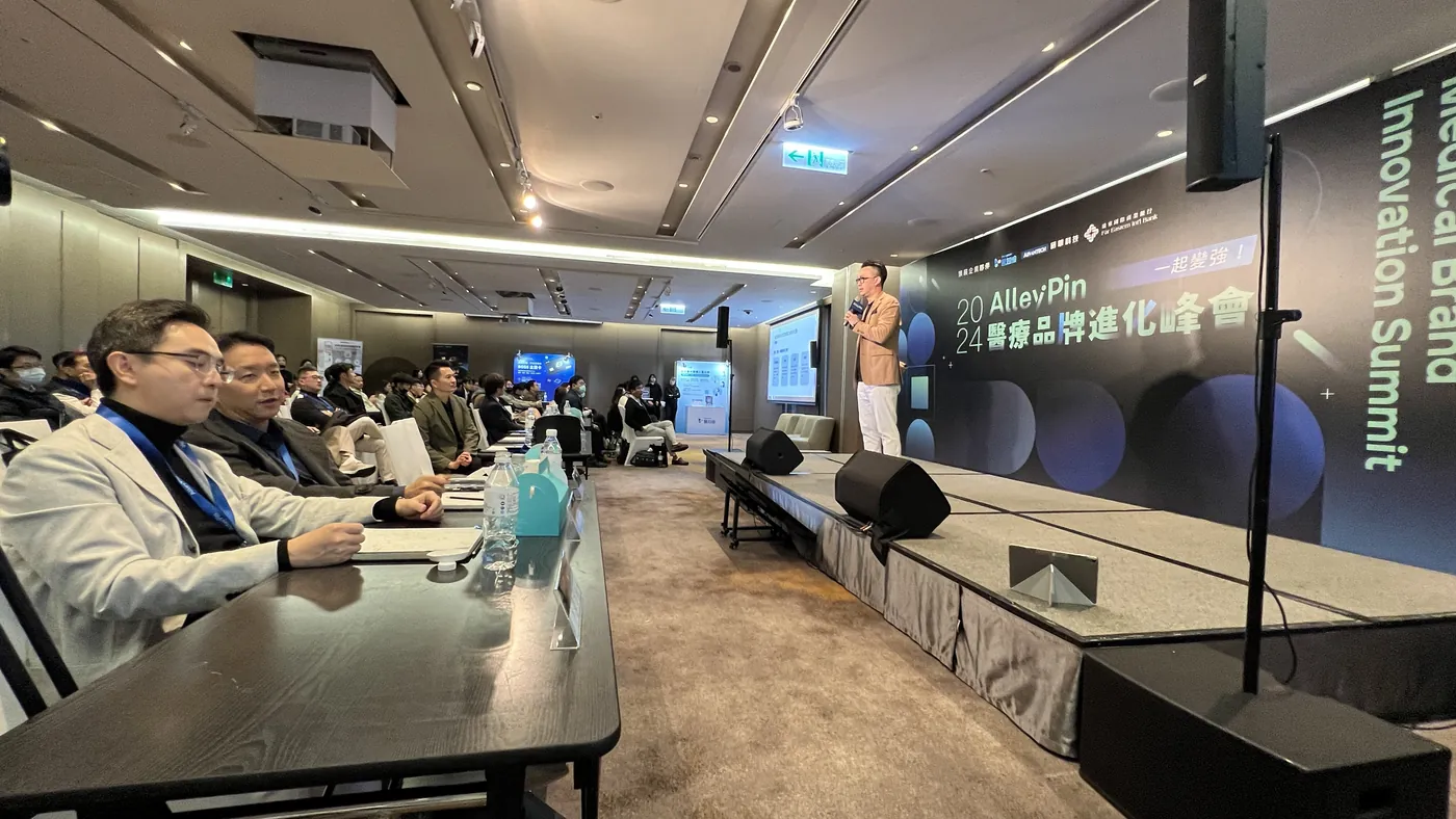 從前排長桌往舞台方向拍：前景是坐在桌旁的聽眾，舞台上有講者正在講話