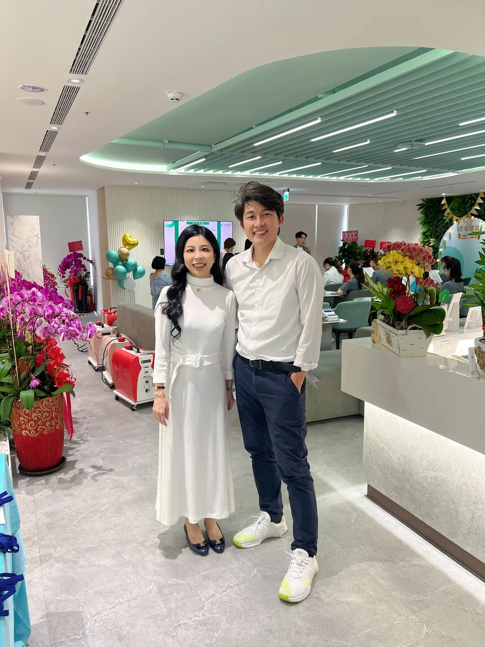 張益豪醫師在康博診所竹北分院大廳與一位白衣來賓合影，身後是成排的祝賀蘭花