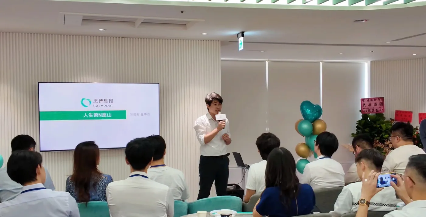 張益豪醫師手持麥克風站在大廳前方開場，旁邊螢幕顯示康博集團簡報首頁「人生第N座山 吳俊毅 董事長」，與會醫師坐在台下