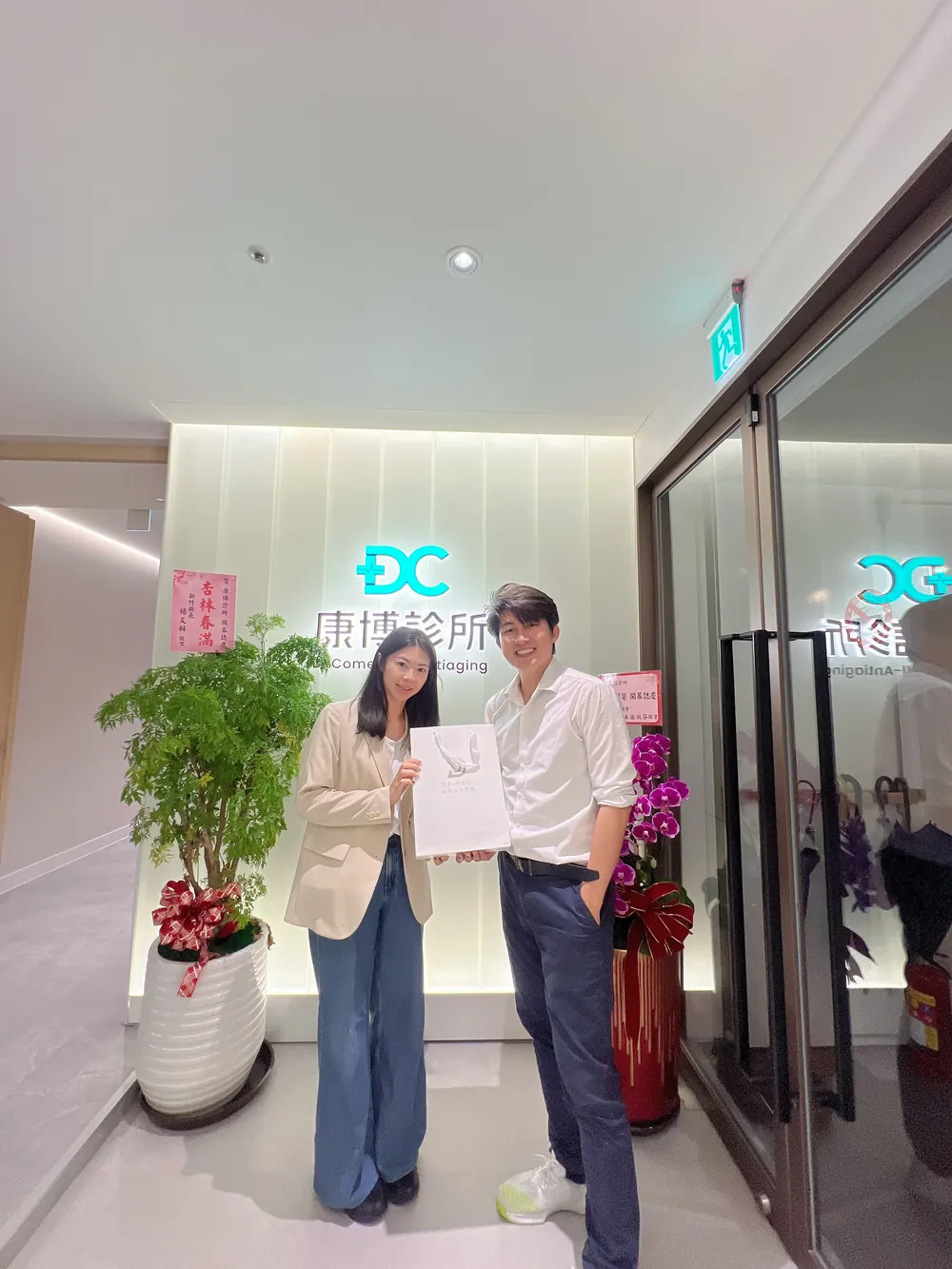 張益豪醫師與一位穿米色西裝外套的女醫師在招牌前拿著伴手禮袋合影