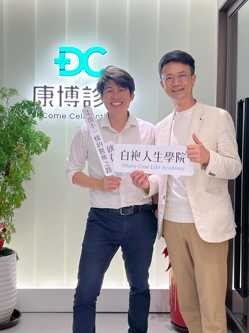 張益豪醫師與一位醫師在康博診所發光招牌前，舉著「白袍人生學院」與「走出不一樣的醫療之路 就在白院」立牌