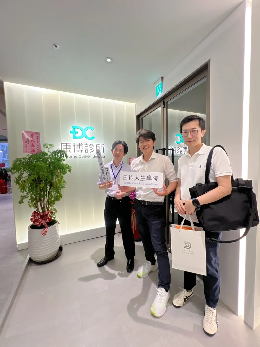 楊為傑醫師、張益豪醫師與徐嘉賢醫師在康博診所招牌前合影，一人舉著白袍人生學院立牌