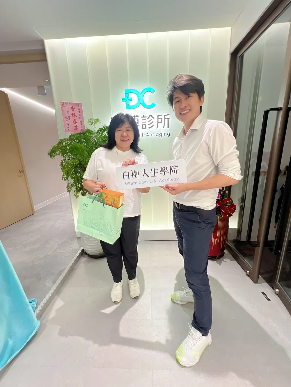 張益豪醫師與一位女醫師在招牌前，一起舉著白袍人生學院立牌，對方手上提著綠色禮袋