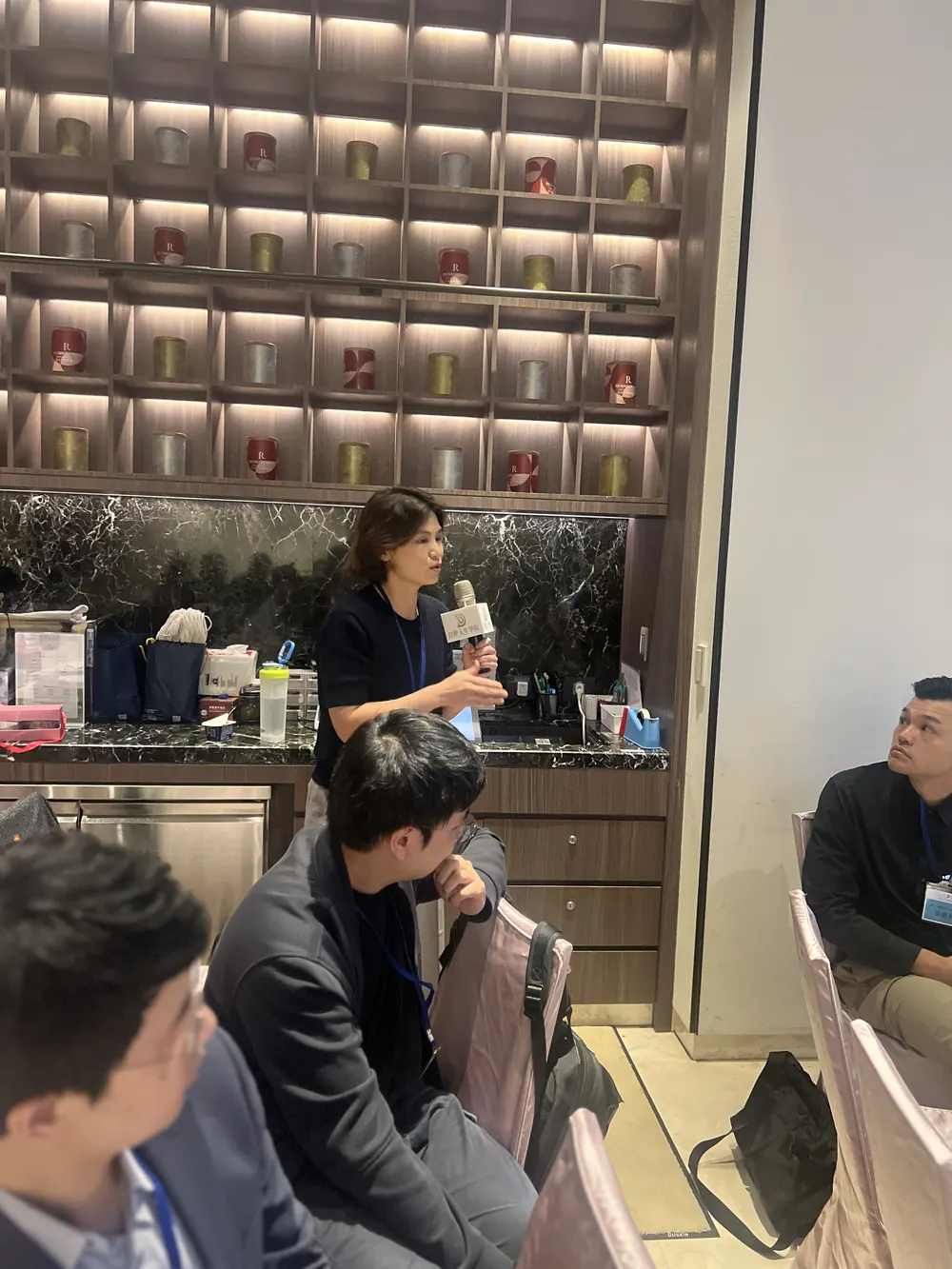 一位與會者在茶廳後方手持麥克風分享，圓桌旁的醫師回頭聆聽
