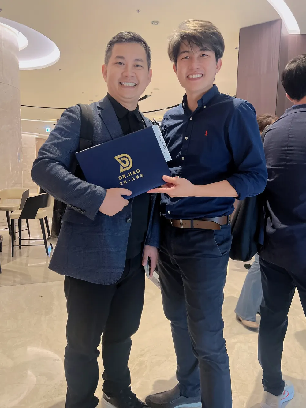 張益豪醫師與一位參訪醫師合影，對方手持印有 DR.HAO 白袍人生學院字樣的深藍色資料夾
