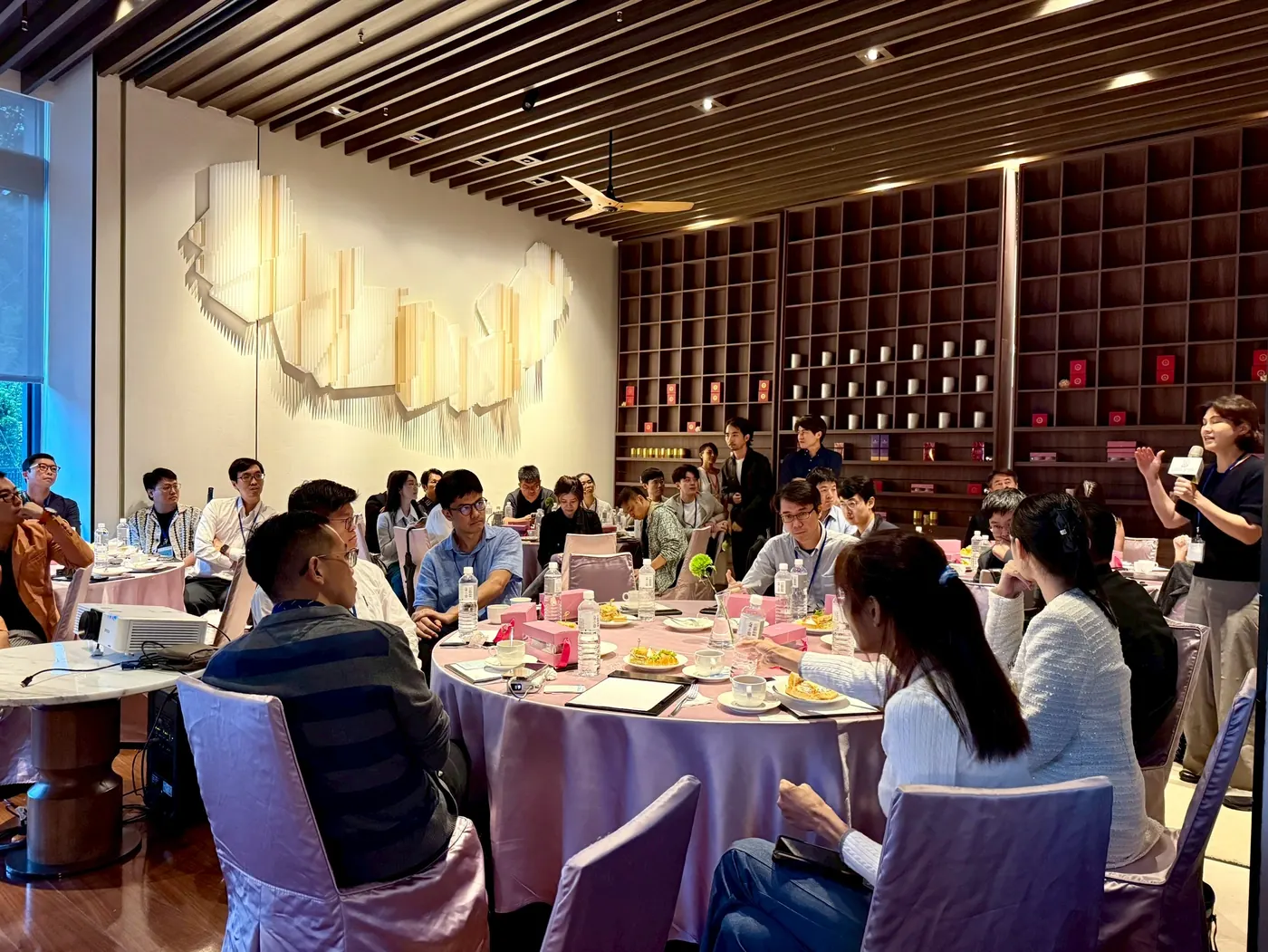 茶廳全景，右側一位與會者手持麥克風發言