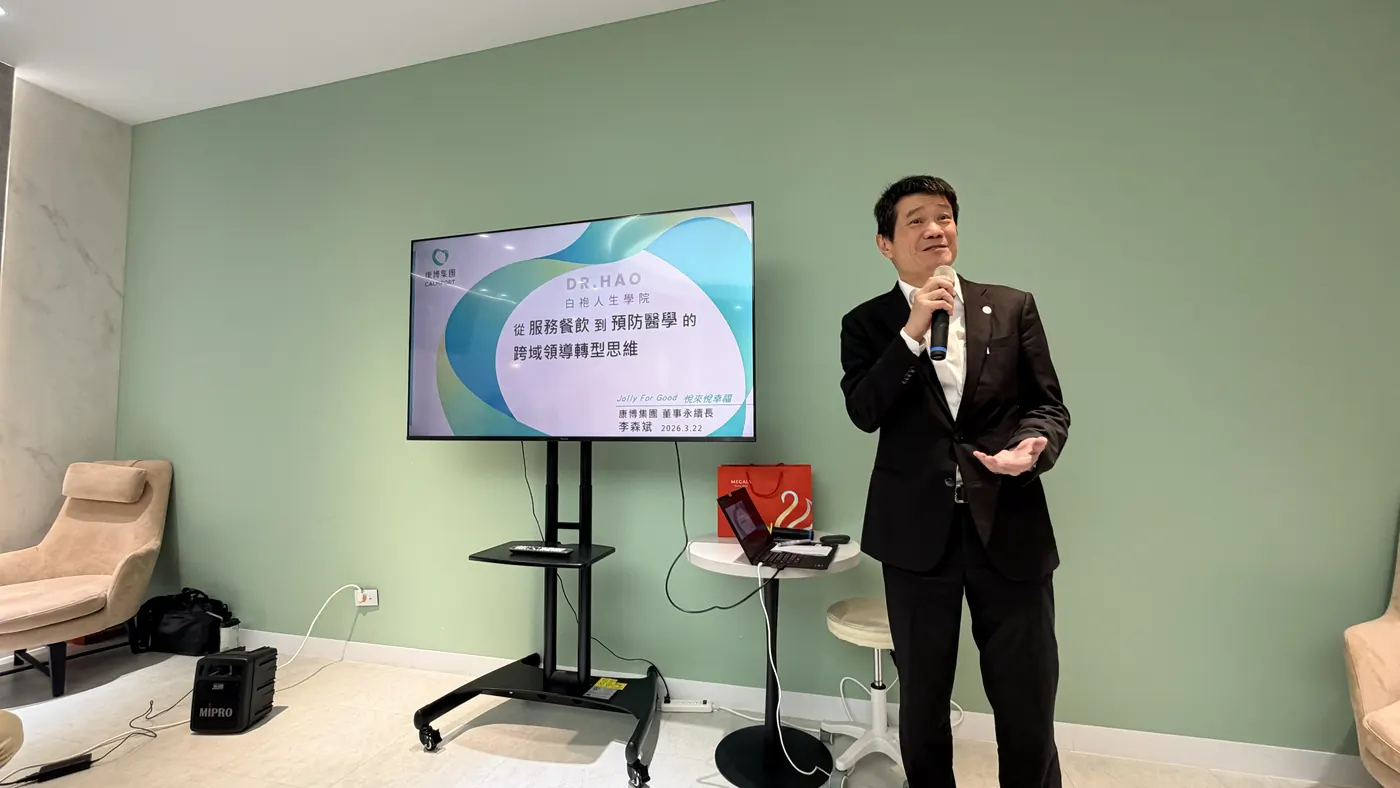 李森斌手持麥克風，站在投影片旁，投影片寫著 DR.HAO 白袍人生學院、從服務餐飲到預防醫學的跨域領導轉型思維、康博集團董事永續長李森斌