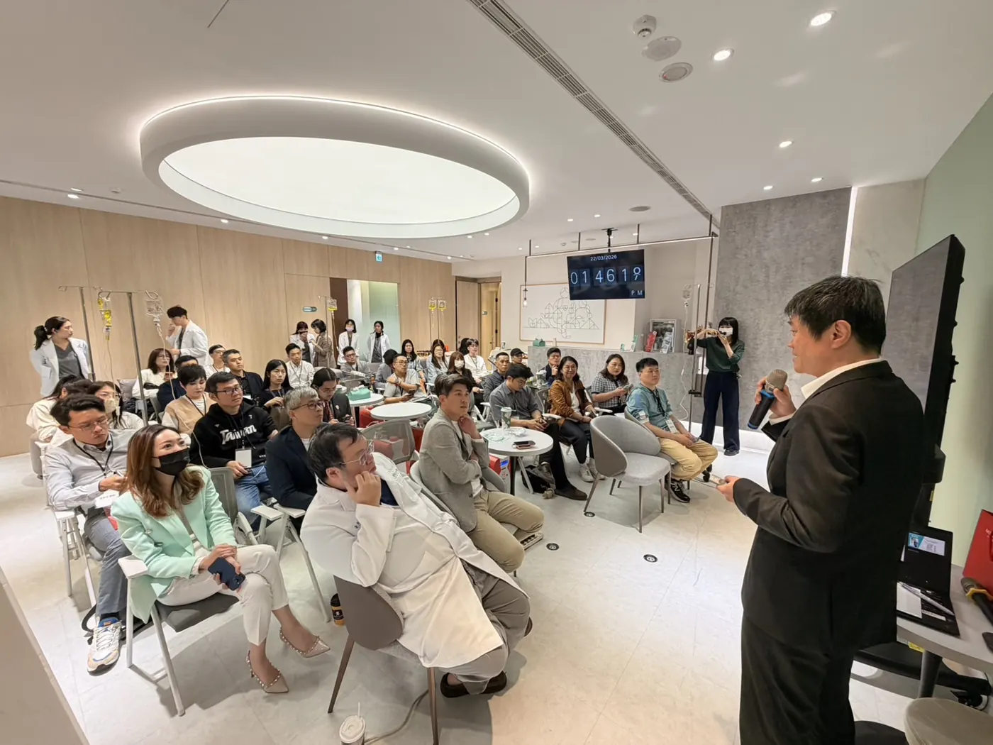 穿西裝的講者站在右側面對全場，台下醫師圍坐聆聽