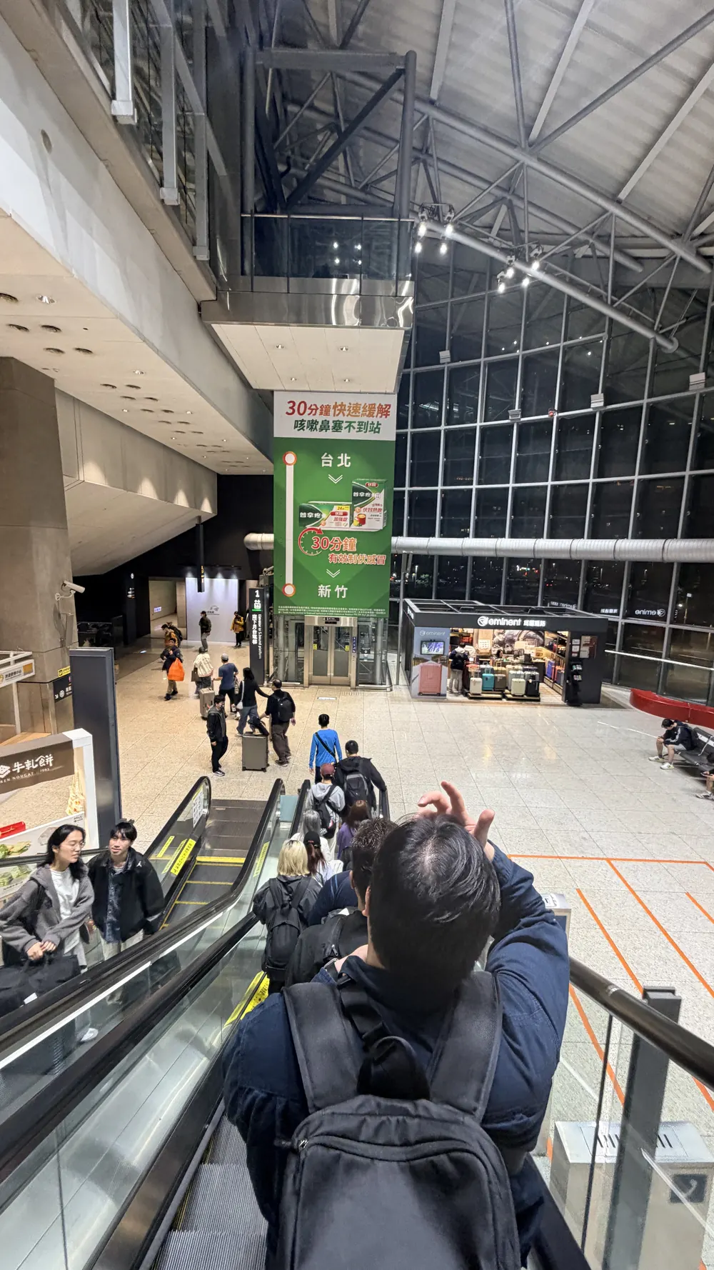 新竹高鐵站大廳，手扶梯往上的方向，遠處掛著一幅綠色的普拿疼伏冒看板