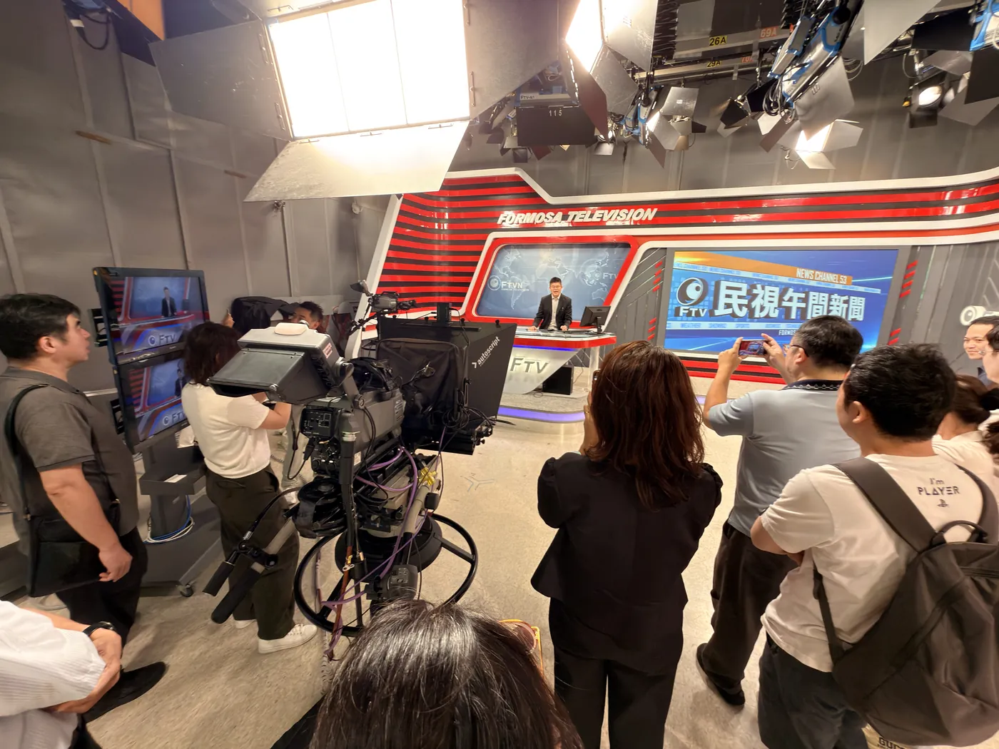 參訪醫師站在民視午間新聞的攝影棚內，圍在攝影機旁看主播台