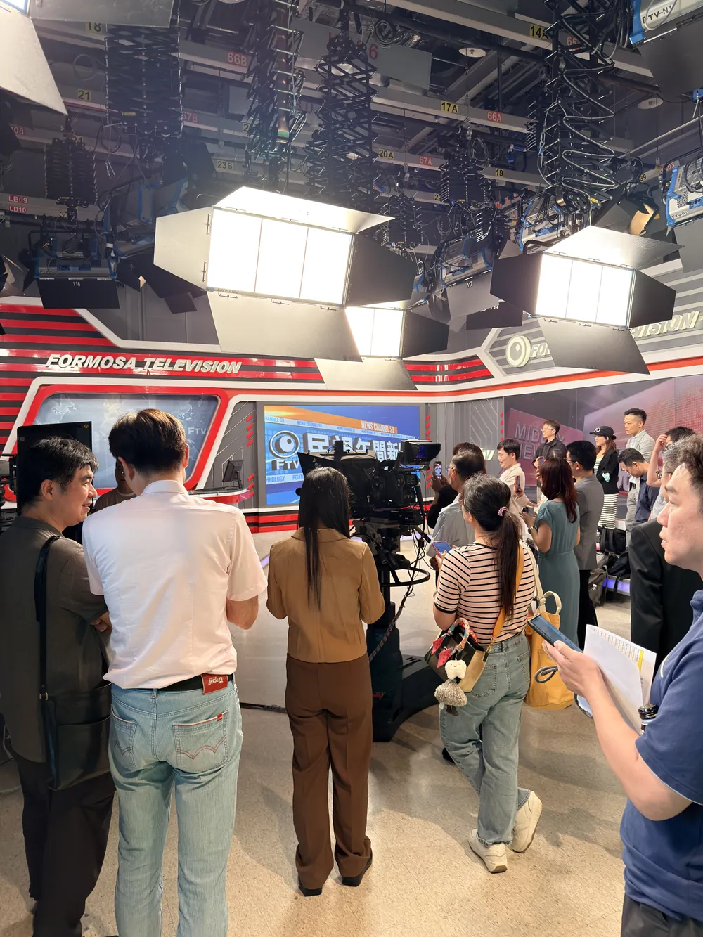 新聞攝影棚內，參訪醫師排隊等著體驗主播台，上方是成排的棚燈