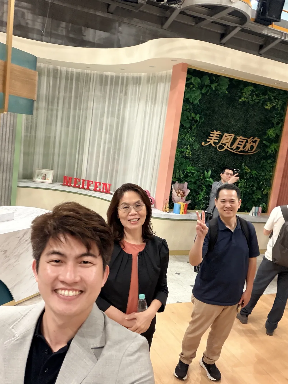 張益豪醫師與林香蘭執行長在《美鳳有約》節目佈景前自拍，後方一位參訪醫師比著手勢