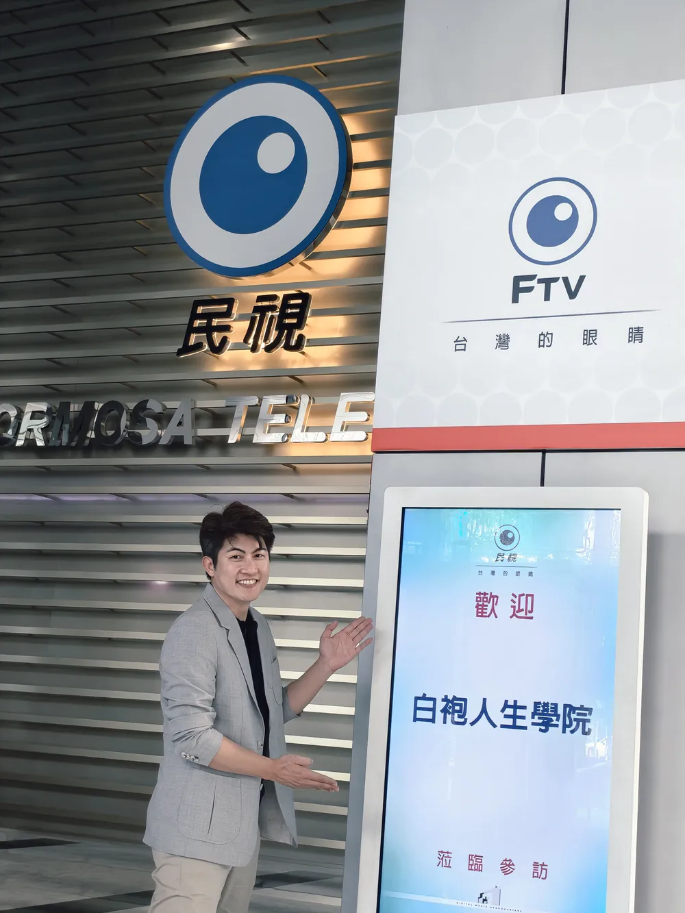 張益豪醫師在民視大廳的歡迎立牌旁比出介紹的手勢，立牌寫著「歡迎 白袍人生學院 蒞臨參訪」