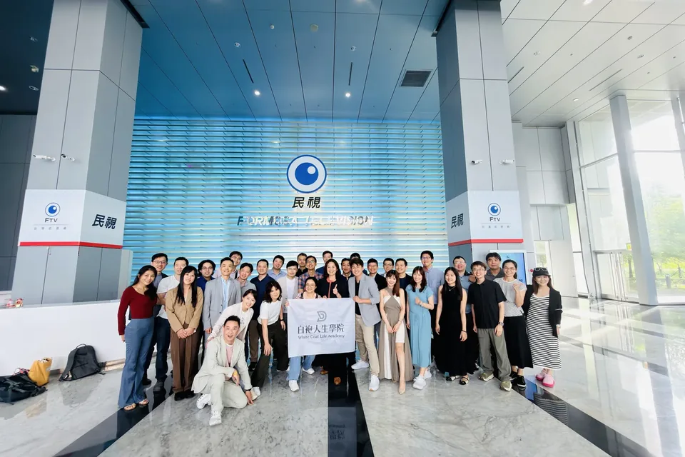 白袍人生學院參訪民視，三四十位醫師在民視一樓大廳的標誌牆前拉著學院旗子合影